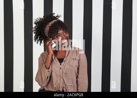 Putting headphone in ear and amazed my the music. Smiled afro american girl stands in the studio with vertical white and black lines at background Stock Photo