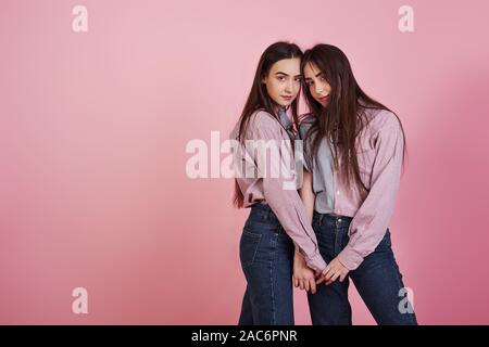 Holding each other by the hands. Young women having fun in the studio with pink background. Adorable twins Stock Photo