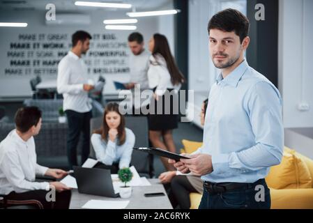 Nice interior. Portrait of young guy stands in the office with employees at background Stock Photo