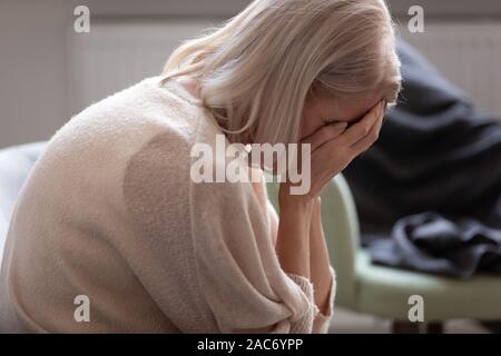 Senior woman hides face with hands crying seated on couch Stock Photo