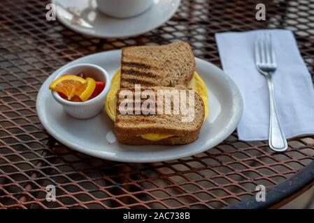Closeup on egg sandwich in wheat bread on white plate inrestaurant setting, outside,  accompanied with warm beverage and fruit - simple breakfast conc Stock Photo