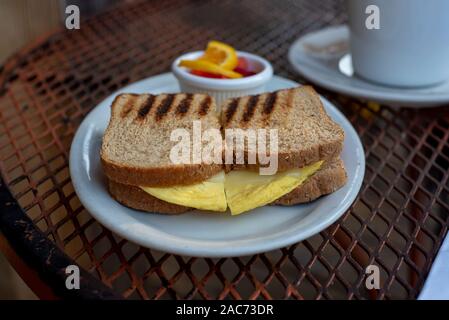 Closeup on egg sandwich in wheat bread on white plate inrestaurant setting, outside,  accompanied with warm beverage and fruit - simple breakfast conc Stock Photo