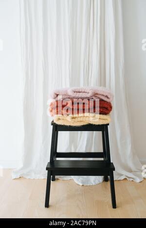 Three folded multi-colored sweaters lie on stepping stool in front of a curtain Stock Photo