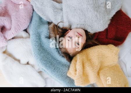 Cute little girl is lying on a pile of soft multi-colored sweaters on the floor. Stock Photo