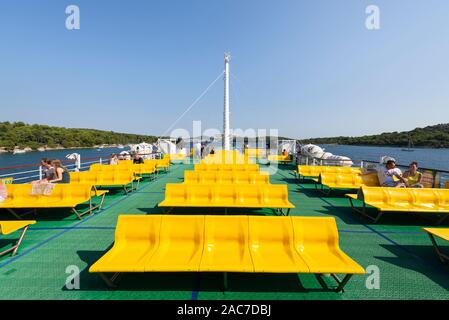 Passengers on a Jadrolinija car ferry in Kvarner Bay in Croatia sit on rows of yellow seats on the green floor of the upper deck in the sun Stock Photo