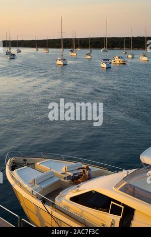 Man and woman sit on the foredeck of a motor yacht in front of anchoring sailing yachts during sunset and drink white wine, Olib, Croatia Stock Photo