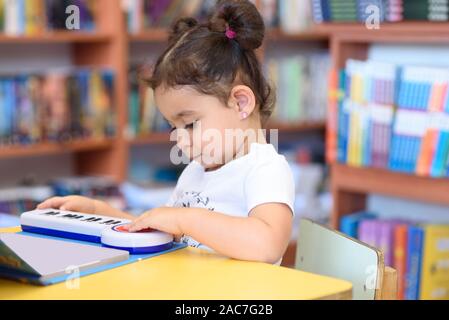 Little Child Indoors In Front Of Books. Cute Young Toddler Sitting On A Chair Near Table and play with toy piano. Kid in a bookstore, surrounded by colorful books. Happy multirace girl play the piano. Stock Photo