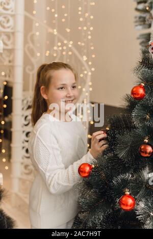 Cute little girl decorates the Christmas tree with New Year's toys and red balls. A girl in a white knitted sweater and dress stands hanging balls on an artificial spruce. Stock Photo
