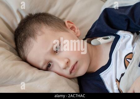 Pre-school sick boy in pyjama lying on pillow in bed with a digital thermometer. Ill boy is measuring body temperature and doesn’t feel well. Stock Photo