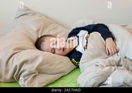 Pre-school sick boy with closed eyes lying on pillow in bed with a digital thermometer. Ill boy is measuring body temperature and doesn’t feel well. Stock Photo