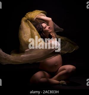 Pregnant woman dancing seated in the dark with a transparent, delicate fabric Stock Photo