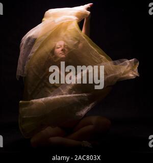 Pregnant woman dancing seated in the dark with a transparent, delicate fabric Stock Photo