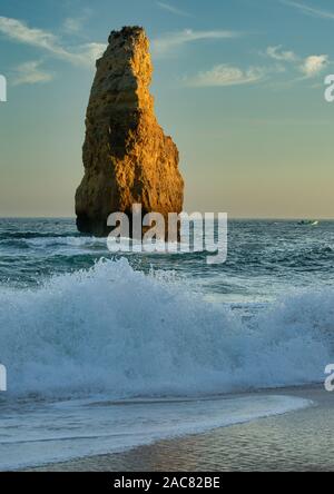 Sandstone cliff island illuminated by evening sun and a wave rushing to the sandy beach of southern Portugal Stock Photo