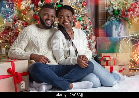 Black couple posing near Christmas tree, sitting on floor and smiling. Stock Photo