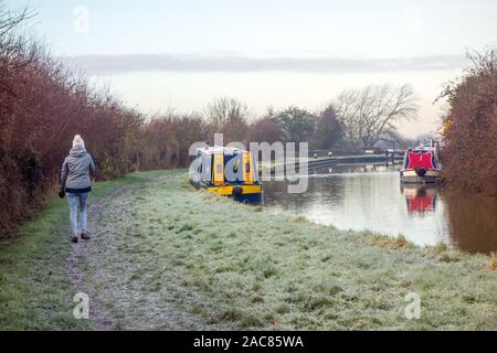 Woman walking along the frosty towpath of the Trent and Mersey canal in the winter with two narrow boats moored up for the day at Hassall Green Stock Photo