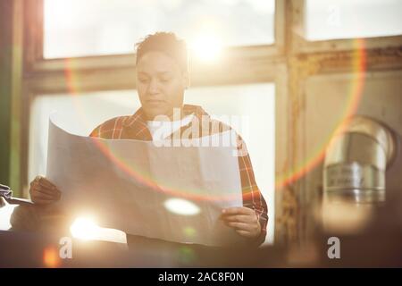 Waist up portrait of mixed-race woman holding engineering plans against sunlit window at industrial workshop, copy space Stock Photo