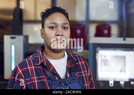 Head and shoulders portrait of mixed-race female worker looking at camera standing by industrial machine units in workshop, copy space Stock Photo