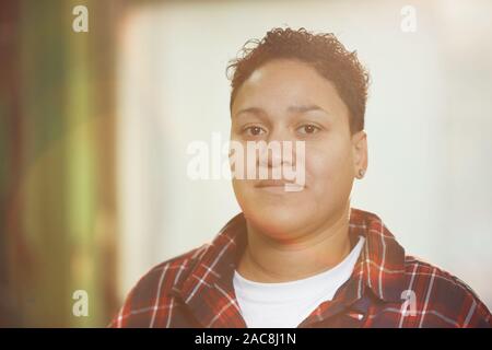 Head and shoulders portrait of modern mixed-race woman working at factory and looking at camera with sun flare, copy space Stock Photo