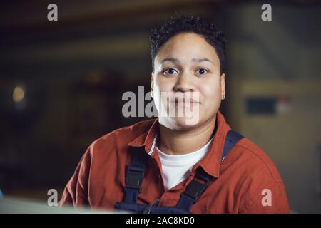 Head and shoulders portrait of female factory worker wearing overalls looking at camera and smiling while posing in industrial workshop, copy space Stock Photo