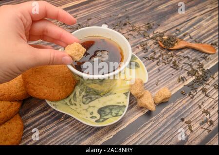 The girl's hand puts a piece of brown sugar in a mug of black tea, oatmeal cookies, tea leaves, brown sugar on a wooden background. Close up Stock Photo