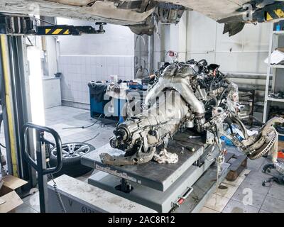 A used car raised on a lift for repair and under it a detached engine and gearbox suspended on a gray lift table near workbench in a vehicle repair sh Stock Photo