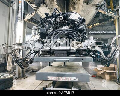 A used car raised on a lift for repair and under it a detached engine and gearbox suspended on a gray lift table near workbench in a vehicle repair sh Stock Photo