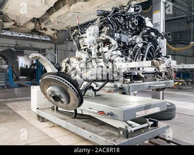 A used car raised on a lift for repair and under it a detached engine and gearbox suspended on a gray lift table near workbench in a vehicle repair sh Stock Photo