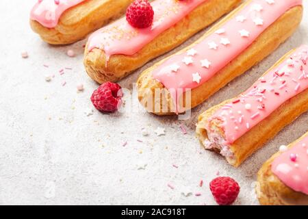 Delicious pink glazed eclairs with cream, raspberry and sugar sprinkles on gray background, copy space. Stock Photo