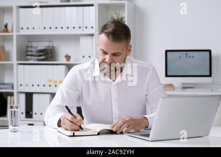 Serious bearded businessman sitting by desk in office, thinking of ideas and making notes in notebook Stock Photo