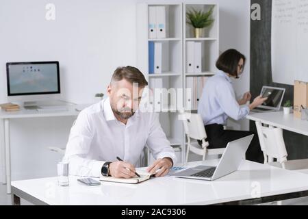 Serious bearded employer thinking of ideas and making notes in notebook on background of colleague using gadgets Stock Photo