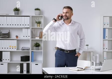 Serious and confident mature entrepreneur in formalwear holding smartphone by his ear while consulting client or business partner Stock Photo