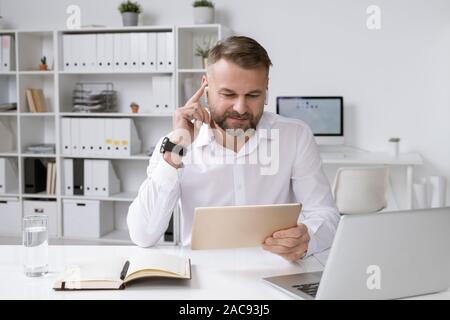 Successful employer with touchpad watching online video while sitting by desk in front of laptop Stock Photo