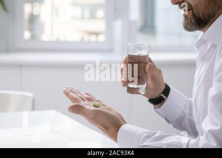 Hands of bearded businessman holding pill while going to take it and glass of water over desk at work Stock Photo