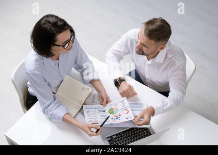 Young female financier and her colleague discussing financial papers at working meeting while sitting by desk Stock Photo