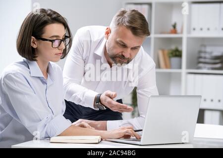 Confident economist pointing at laptop display while showing online information to colleague at meeting Stock Photo