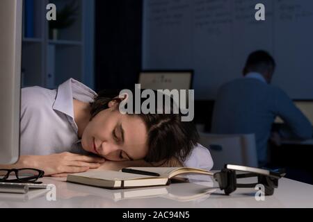 Exhausted young female hotline operator napping with her head on desk after working late in the evening with colleague on background Stock Photo