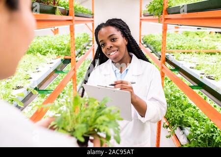 Happy young woman of African ethnicity making notes in document while communicating with colleague in greenhouse Stock Photo