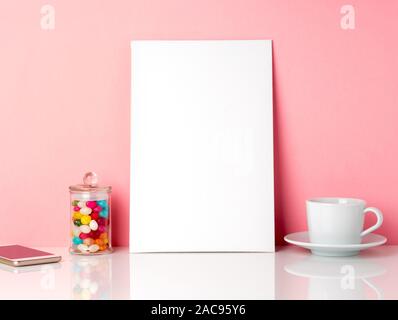 Blank white frame and candys in jar, cup of coffee or tea on a white table against the pink wall with copy space Stock Photo