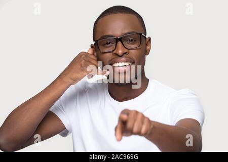 Head shot portrait of smiling African American man in glasses showing call me gesture, looking, pointing finger at camera, happy male with phone shape Stock Photo