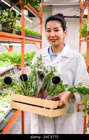 Happy young Asian greenhouse worker in whitecoat carrying organic food Stock Photo
