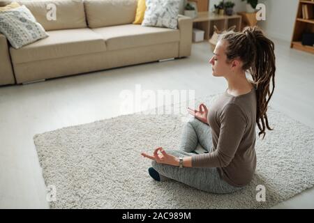 Girl in activewear listening sitting in pose of lotus on carpet while meditating Stock Photo