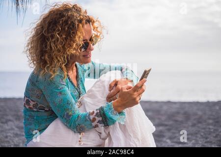 Nice cheerful caucasian woman with boho trendy clothes use modern technology cellular phone sit down at the beach in outdoor leisure activity - people Stock Photo