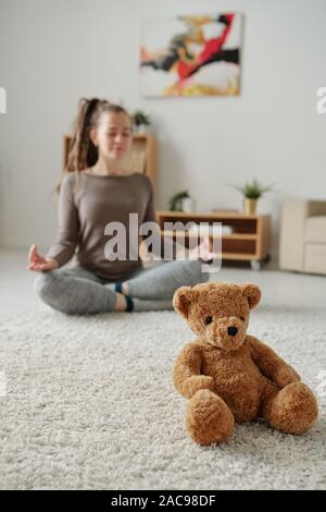 Cute teddybear sitting on carpet with young meditating woman on background Stock Photo