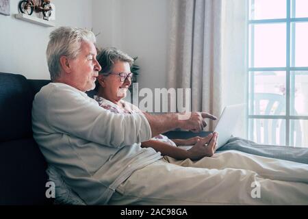 Cheerful happy beautiful caucasian old aged senior couple sit down and relax on the soa at home looking and using a personal computer laptop to enjoy Stock Photo