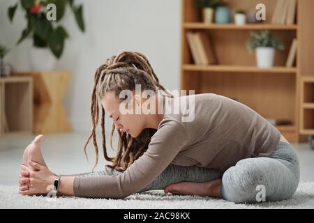 Young flexible woman in activewear sitting on the floor and stretching one leg Stock Photo