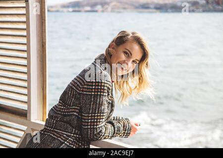 A young Caucasian girl looks out of a window that overlooks the sea and the wind waves her hair, the model is smiling at the camera. Happy travel and vacation concept. Stock Photo