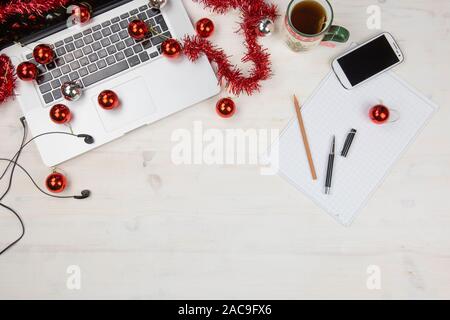 Job at Christmas holidays: flat lay of a light wooden table with an open aluminum laptop, red decoration, red baubles, Christmas cup of tea, smartphon Stock Photo