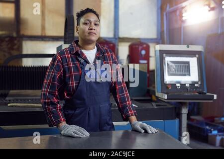 Waist up portrait of mixed-race female worker wearing overalls and looking at camera while standing by industrial machine units in workshop, copy space Stock Photo
