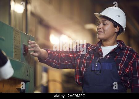 Waist up portrait of smiling female worker pushing red button while operating machine units at production plant, copy space Stock Photo