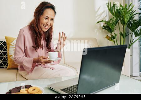 Laughing young woman with cup of coffee waving with hand when greeting friend video calling her Stock Photo
